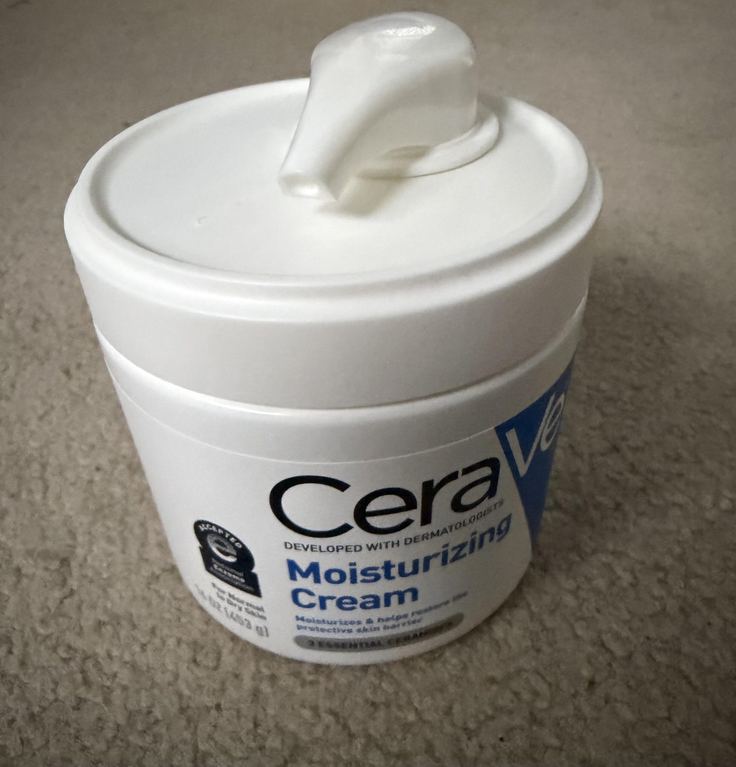 Open CeraVe Moisturizing Cream tub with a dollop of the whippy cream on a glass surface