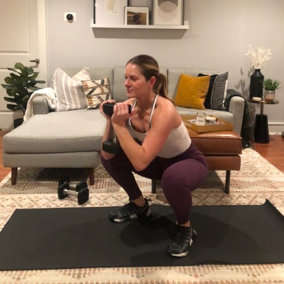 A plus-size pregnant woman doing a goblet squat to a chair at home, holding a small dumbbell