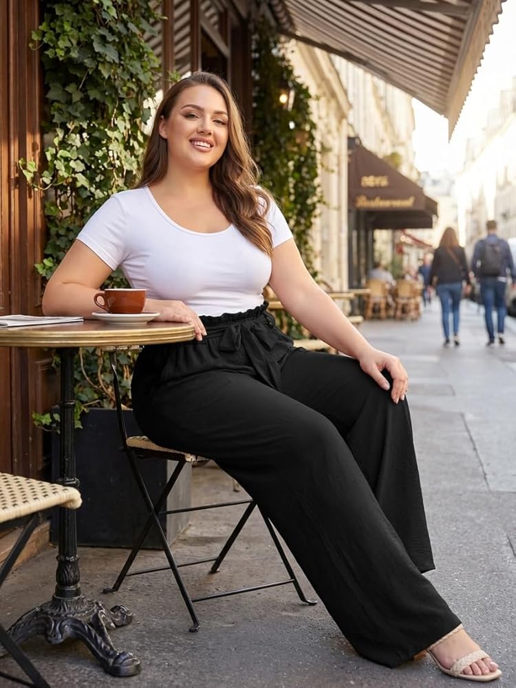 A plus-size woman wearing a wide-leg trouser and a tucked white tee standing in a sunlit modern apartment